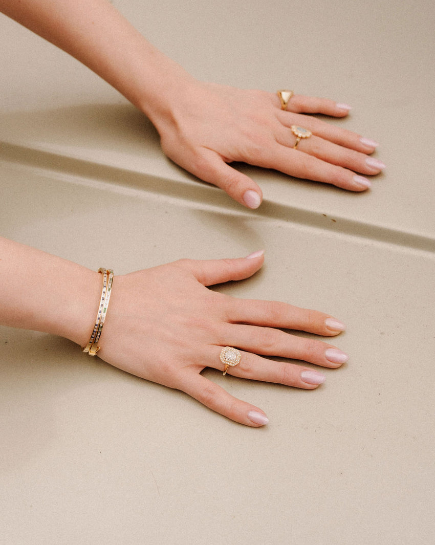 Two hands wearing gold rings and bracelets on a beige surface